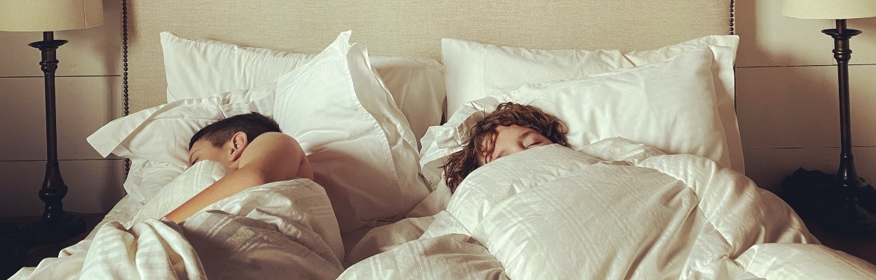 Two people sleeping in a queen bed with white sheets and big white pillows.