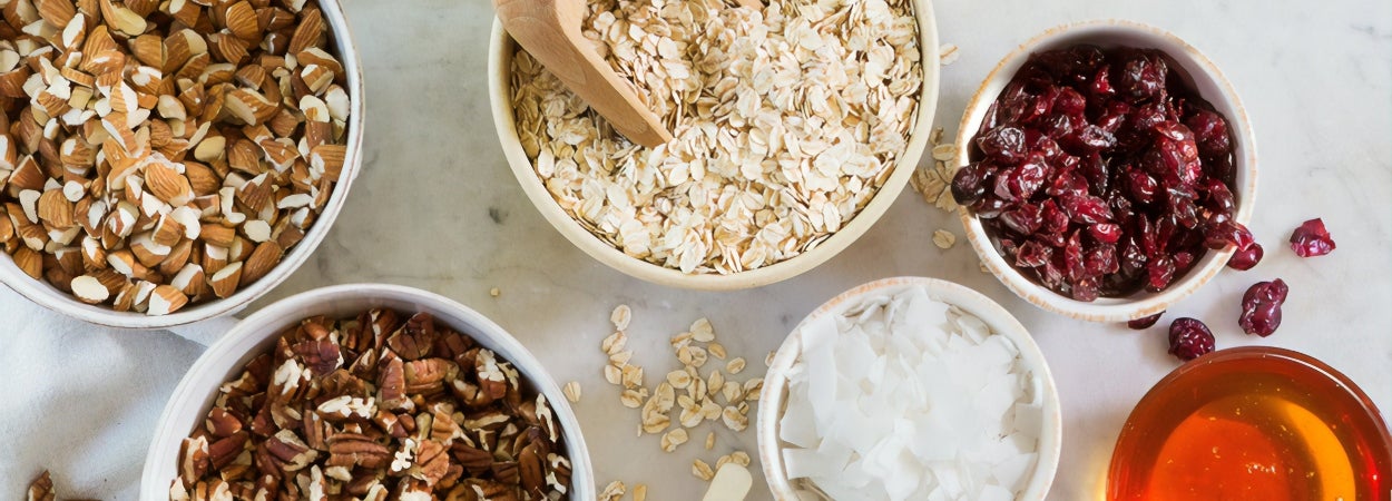 Bowls of protein ball recipe ingredients on a white bench top including honey, oats, nuts and dried fruit.